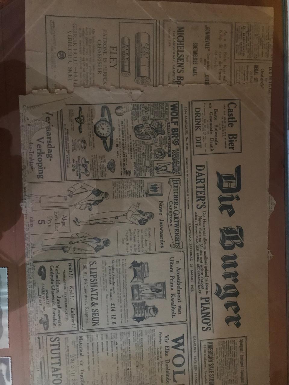 A historical newspaper titled 'Die Burger' featuring advertisements and articles, displayed in a frame.