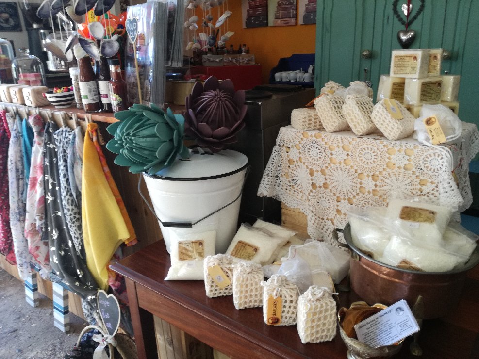 A cozy display of Ouma Hanna se Boerseep products, featuring traditional cold-water soaps, packaged bars, and grated soap arranged on wooden shelves and tables, with colorful scarves as a backdrop and decorative elements like succulents.