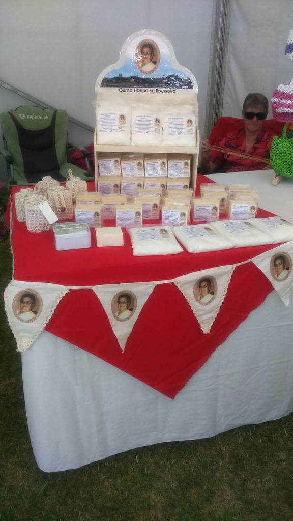 A display of Ouma Hanna se Boerseep products on a table, featuring various soaps packaged in bags and boxes. The setup includes a banner with the brand name and images associated with the product, set against a red tablecloth.
