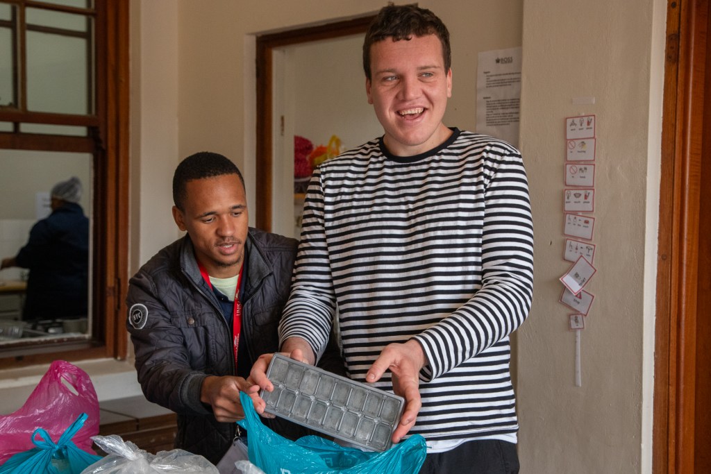 Two individuals engaged in a supportive activity at the Bonnievale Autism Care Centre, one holding an ice cube tray while the other assists.