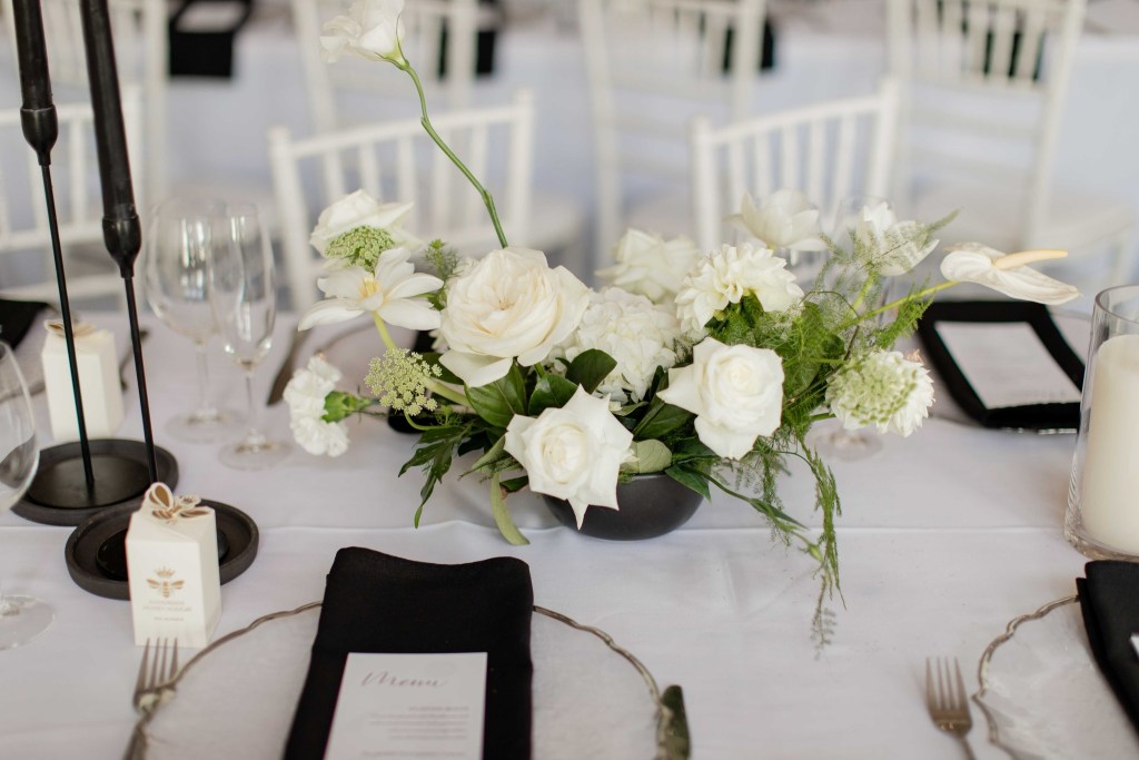 A beautifully arranged centerpiece of white flowers, including roses and lilies, set on a dining table with elegant tableware and a menu displayed.
