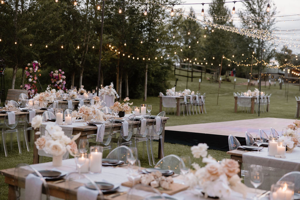 Outdoor event setup with elegantly decorated tables featuring floral centerpieces and candlelight, string lights overhead creating a charming atmosphere.