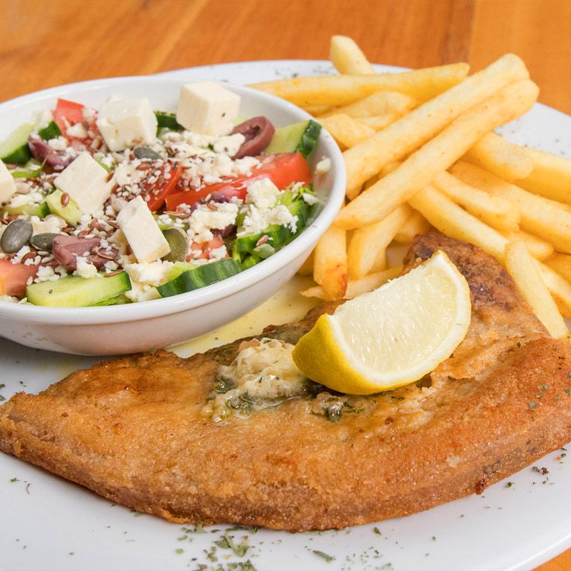 A plate featuring a golden fried fish fillet, accompanied by a side of crispy French fries and a fresh salad topped with feta, olives, and diced vegetables, garnished with a lemon wedge.