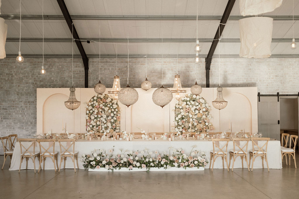 Elegant event setup featuring a long table adorned with floral arrangements and candles, surrounded by wooden chairs, with stylish chandeliers and a floral backdrop in a spacious venue.