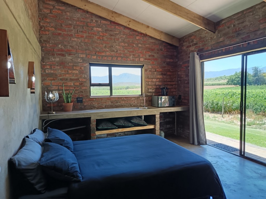 Interior of a guest unit at Merwespont, featuring a brick wall, a double bed with dark blue bedding, and a kitchenette with a microwave. Large windows offer a view of vineyards and mountains outside.