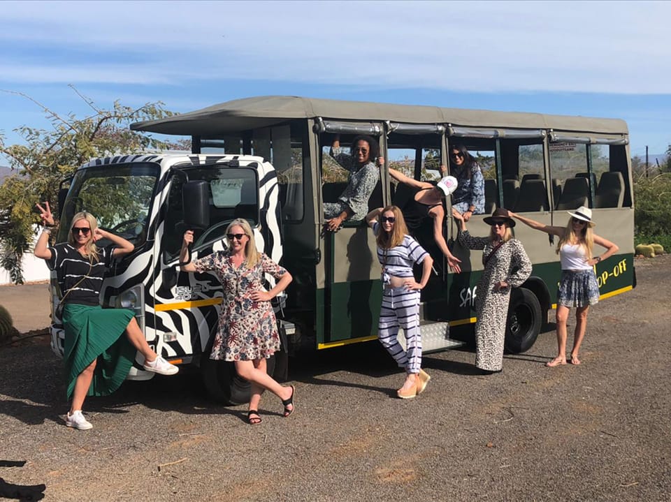 Group of people posing joyfully around a zebra-striped safari vehicle in a scenic outdoor setting.
