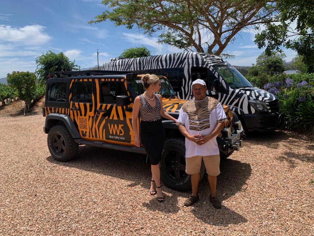 A woman in a black dress stands next to a man wearing a patterned shirt, both posing in front of a zebra-striped Jeep and a matching safari vehicle in a vineyard setting.