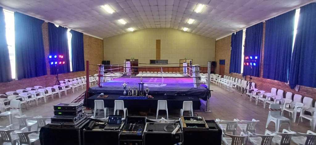 Interior view of a boxing ring set up in a spacious hall with white chairs arranged around it and lighting equipment in the background.