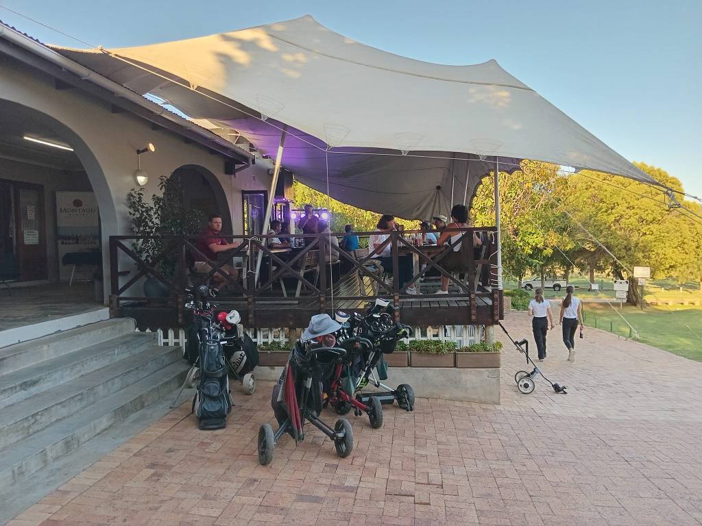 Outdoor seating area with a large canopy, where people are enjoying drinks and food, overlooking a green space. Golf bags and carts are parked nearby.
