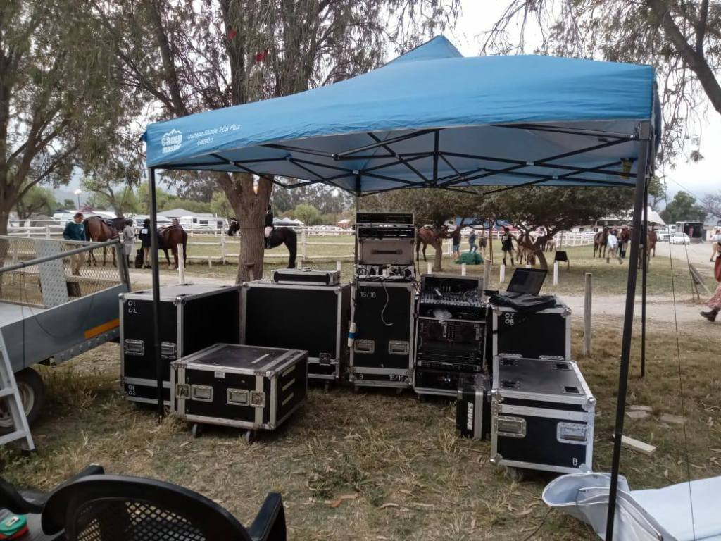 A blue canopy tent set up at an outdoor event, surrounded by various audio equipment cases and horses in the background.