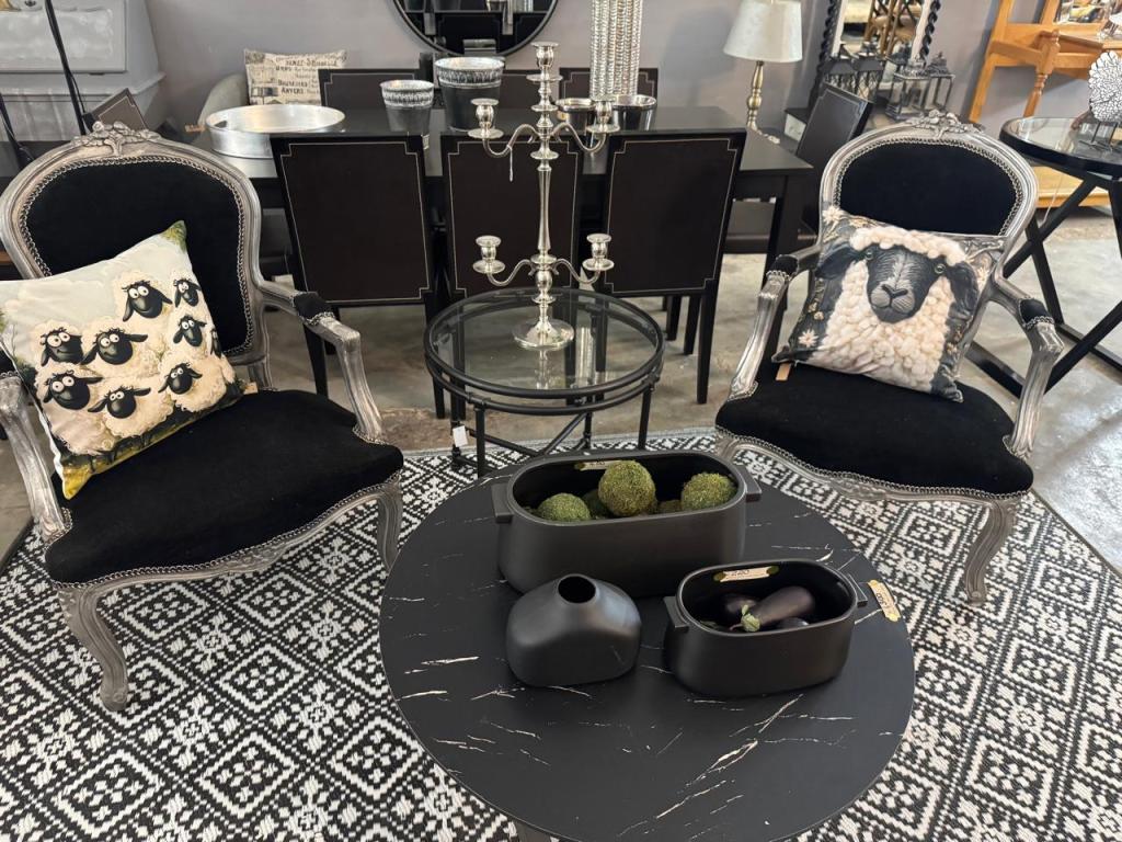 Two elegant black chairs with decorative pillows, one featuring sheep and the other a sheep face, arranged around a round coffee table. The table holds minimalist black decor items and is set on a patterned rug.