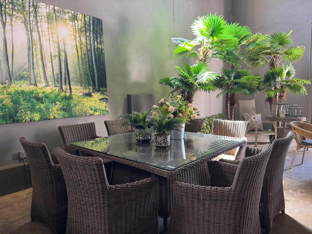 A modern dining area featuring a large glass-top table surrounded by woven chairs. Lush green plants and a floral arrangement are present on the table. A large nature-themed artwork depicting a forest with sunlight filtering through trees hangs on the wall.