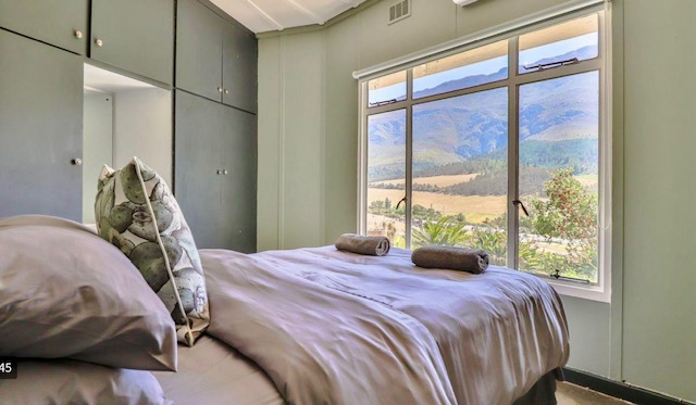 Cozy bedroom featuring a bed with gray linens and decorative pillows, windows offering views of mountains and lush greenery.