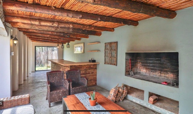 A rustic outdoor living space featuring a wooden ceiling, a bar area, wicker chairs, a wooden table with a potted plant, and a brick fireplace.
