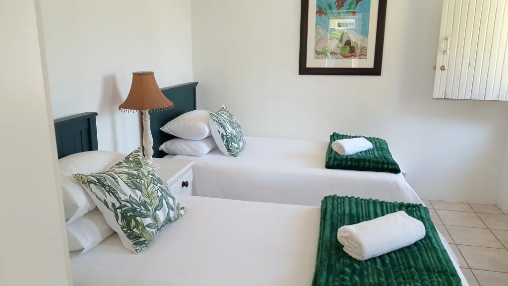 A cozy and well-decorated bedroom featuring two single beds with green throws and white towels, a bedside lamp, and a framed piece of artwork on the wall.