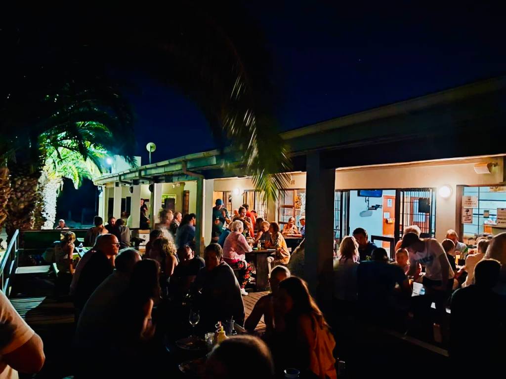 A vibrant outdoor dining scene at night with numerous people enjoying food and drinks, surrounded by palm trees and illuminated by soft lighting.