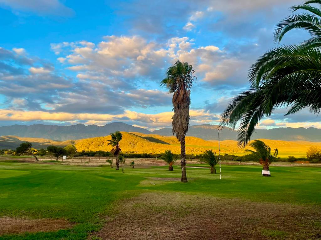 Scenic view of a golf course featuring green lawns, palm trees, and mountains under a blue sky with scattered clouds.