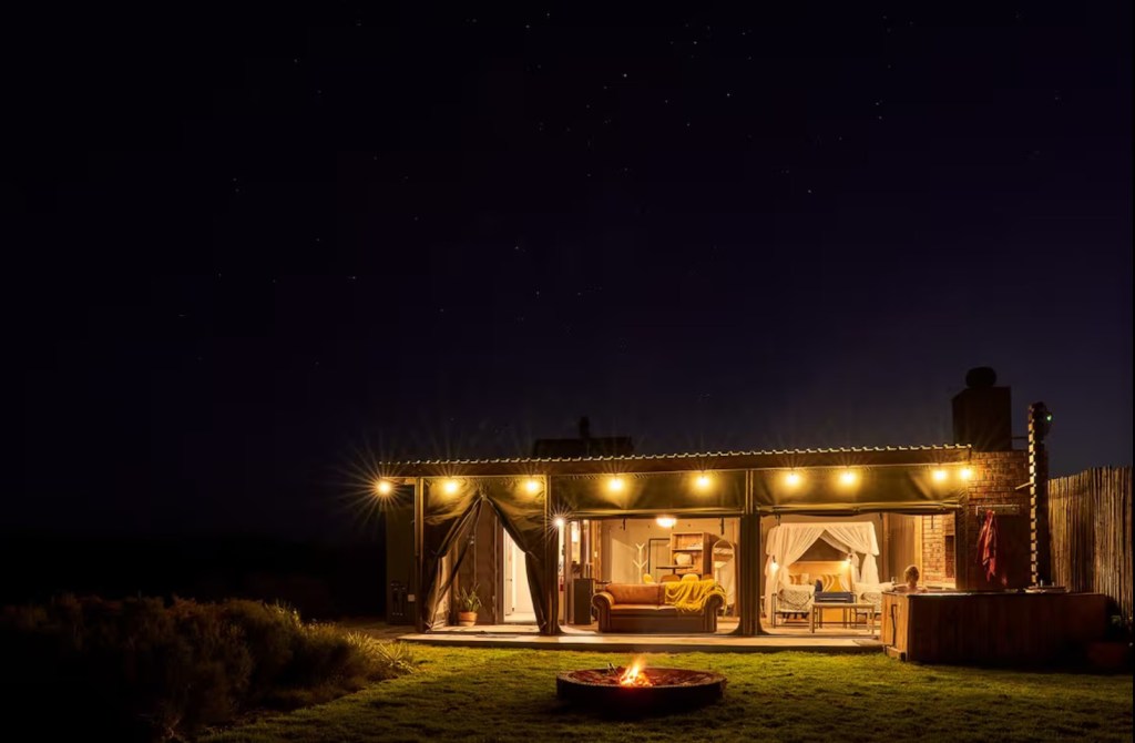 Cozy outdoor view of a modern cabin at night, featuring warm lights, a fire pit, and comfortable seating visible through large openings.