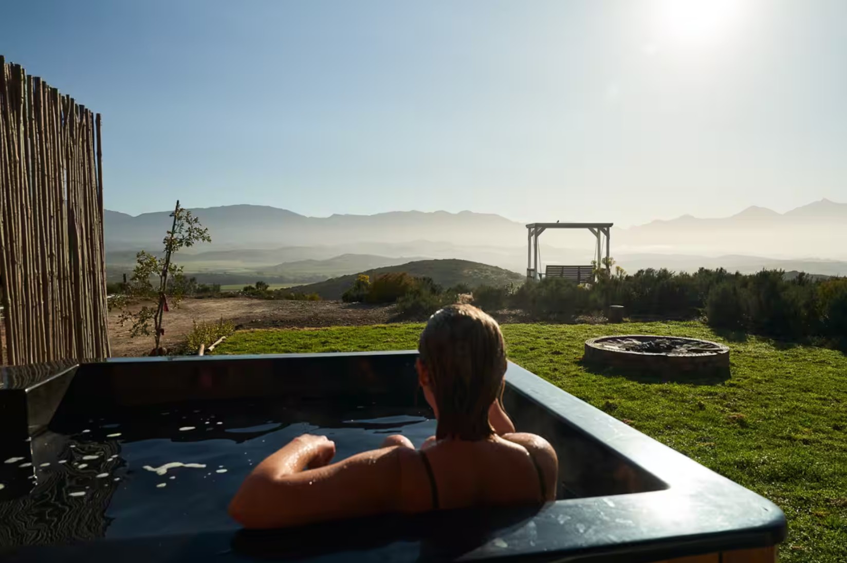 A person relaxing in a hot tub overlooking a scenic landscape with mountains and a clear sky.