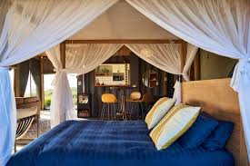 A cozy bedroom featuring a wooden bed with blue bedding and decorative pillows, surrounded by sheer white curtains. The room has a modern aesthetic with a view of a small kitchenette in the background.