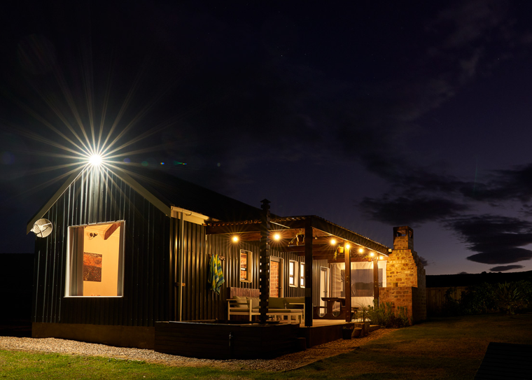 A modern black-clad house at night, illuminated by external lights, with a wooden deck and chimney.