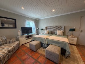 A modern guest room featuring two single beds with plush headboards, a television, a striped sofa, and decorative ottomans.